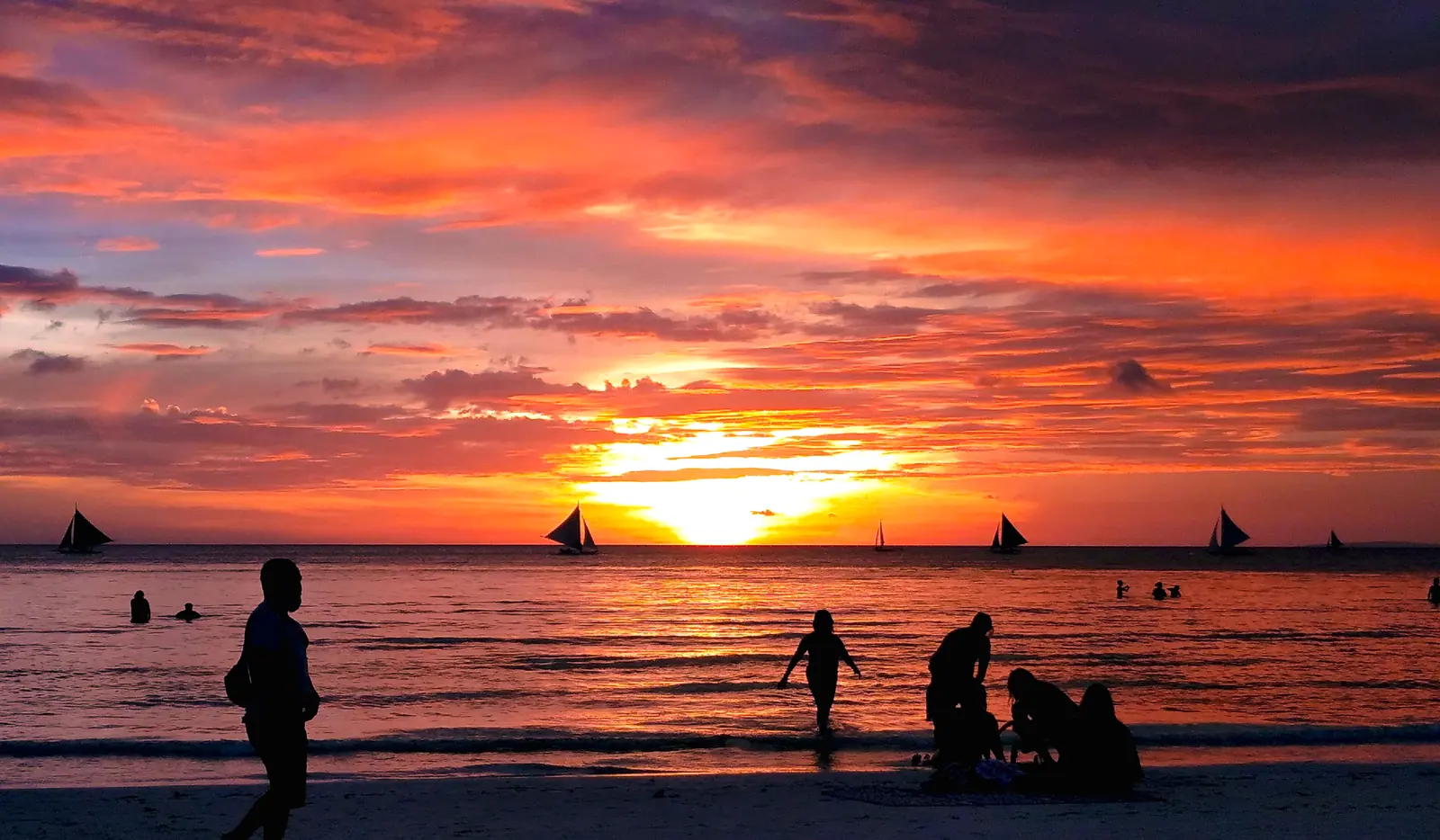 Sunset over White Beach, Boracay: paraw sailboats on the water and people at the shoreline
