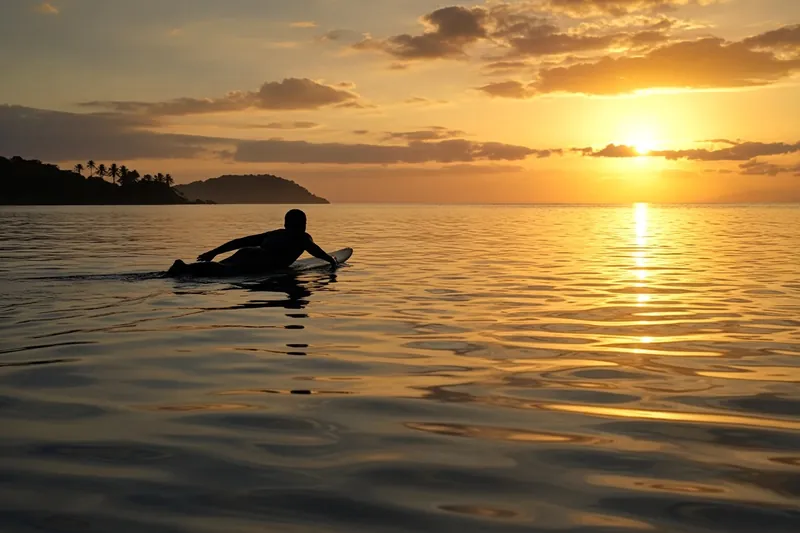 A surfer paddling out across glassy water in golden light