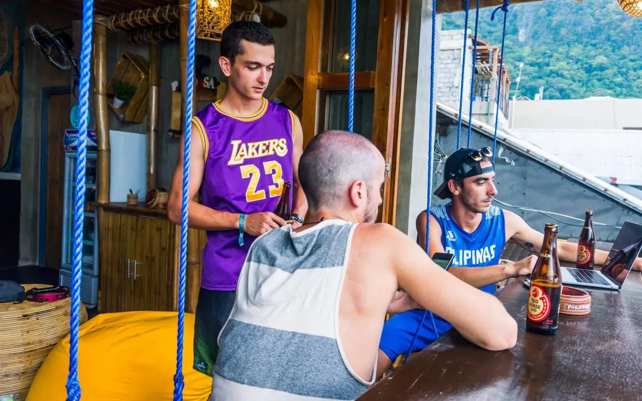 Guests sharing a beer at the Happiness Hostel swing bar, with El Nido Town's green hillside behind